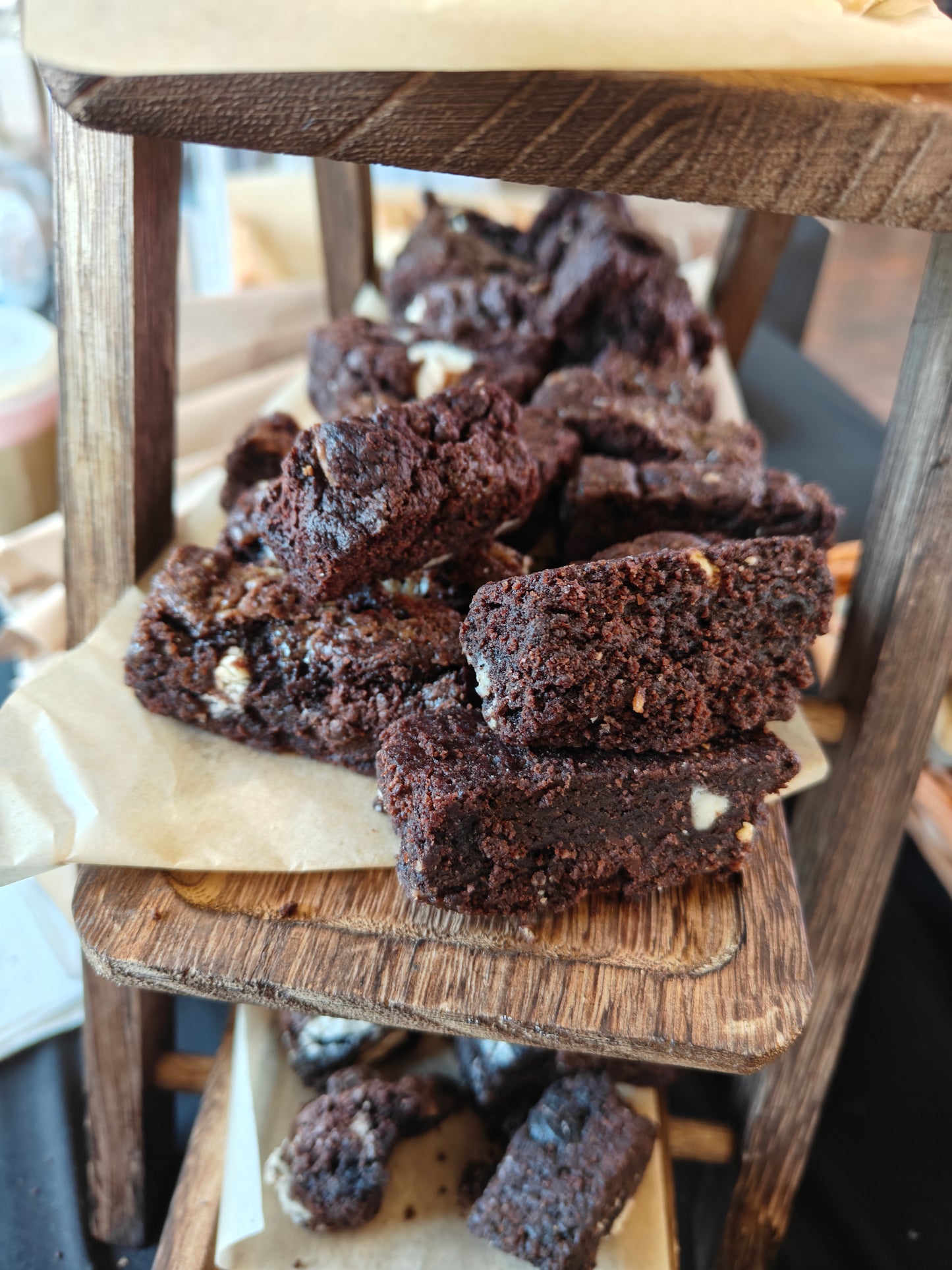 Bite-sized pieces of triple chocolate brownie with white chocolate, dark chocolate, and crushed pretzel on a wooden chopping board with a farmers market in the background