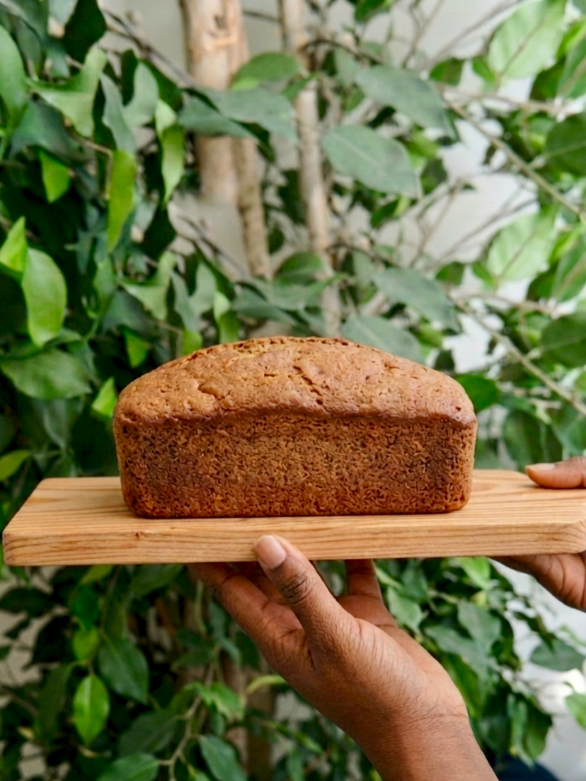 Coffee loaf cake on a wooden board held by two hands against a green foliage background
