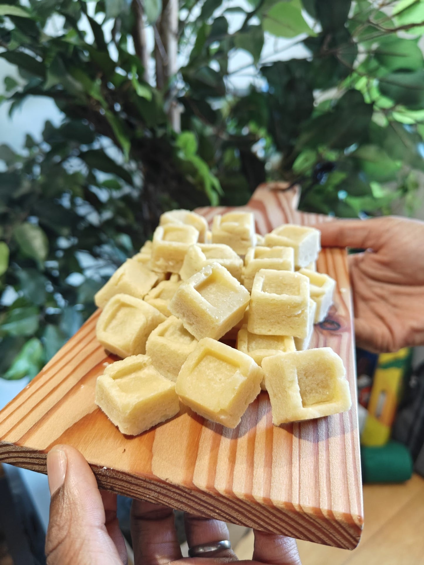 A close-up of small pale yellow square shortbread piled on a wooden board, held by hands in front of a blurred background of green indoor plants.