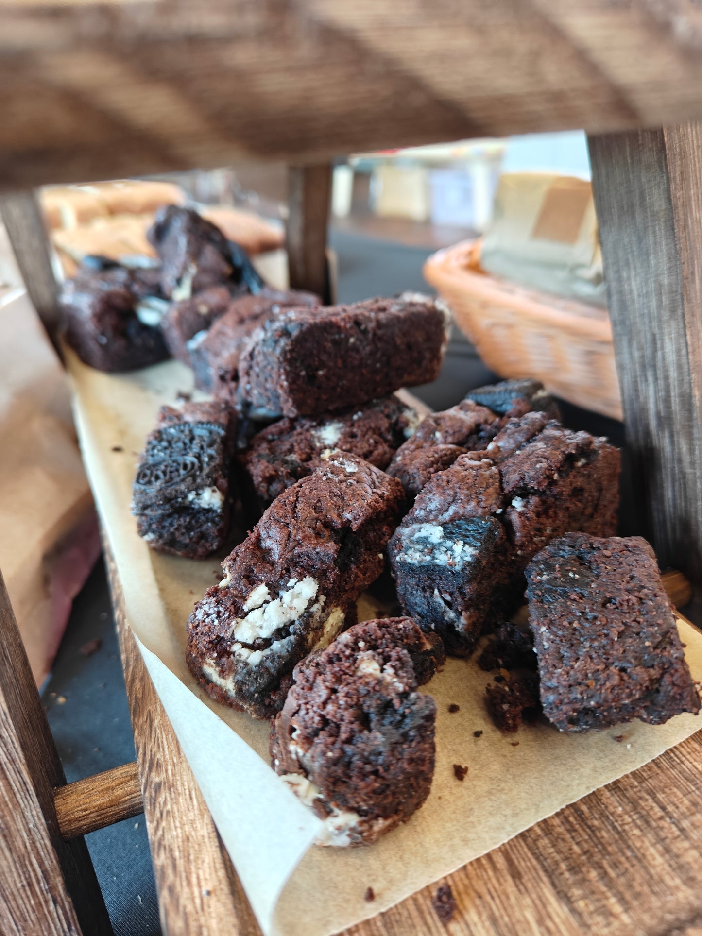 Bite-sized pieces of triple chocolate brownie with white chocolate, dark chocolate, and crushed Oreo on a wooden chopping board with a farmers market in the background