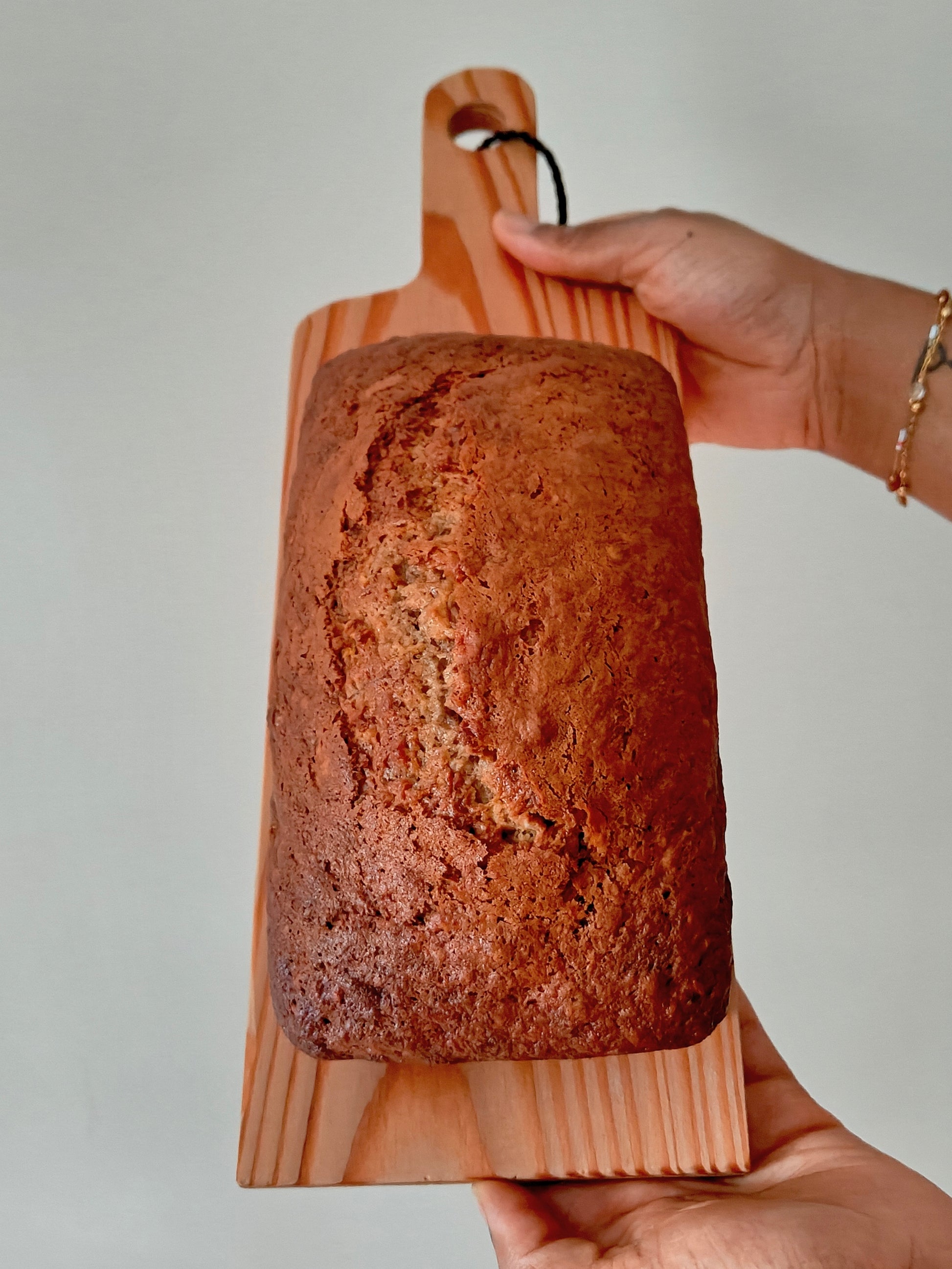Loaf of banana bread on a wooden board held by two hands against a neutral background