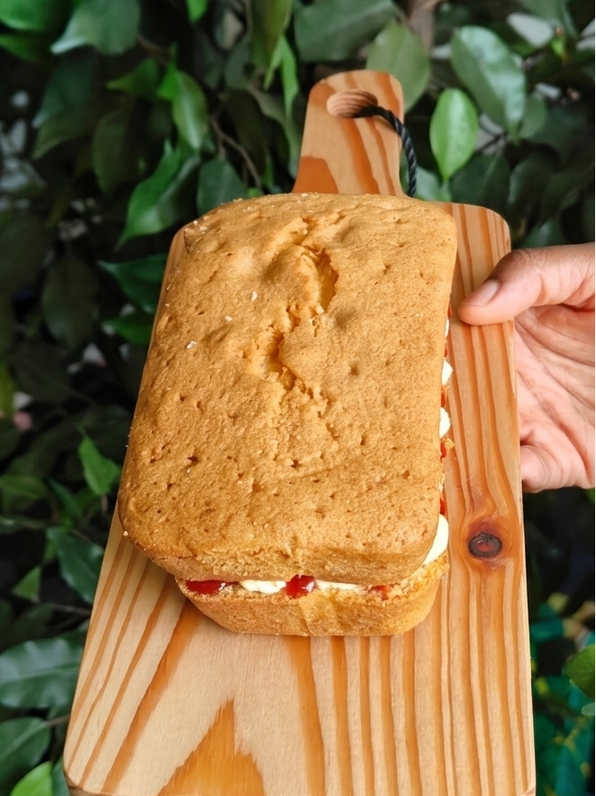 Victoria Sponge Loaf cake with mascarpone buttercream filling and bonne maman strawberry jam on a wooden board, held by a hand with a blurred foliage background.