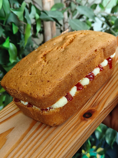 Victoria Sponge Loaf cake with mascarpone buttercream filling and bonne maman strawberry jam on a wooden board, held by two hands with a blurred foliage background.