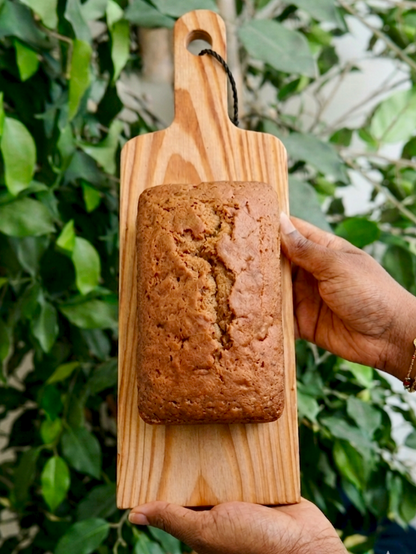 Coffee loaf cake on a wooden board held by two hands against a green foliage background