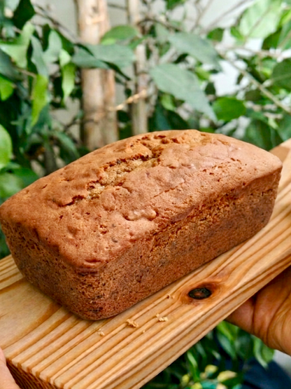 Coffee loaf cake on a wooden board held by two hands against a green foliage background
