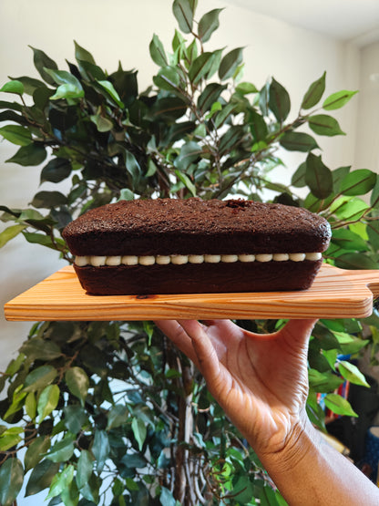 A chocolate loaf cake with white mascarpone buttercream frosting icing on a wooden board, held by two hands in front of a green potted plant and a bookshelf.