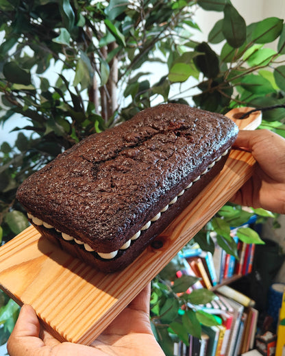 A chocolate loaf cake with white mascarpone buttercream frosting icing on a wooden board, held by two hands in front of a green potted plant and a bookshelf.